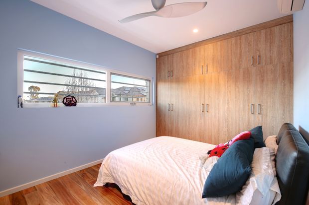 Compact bedroom with a large built-in timber wardrobe spanning one wall, warm timber flooring, pale blue walls, and a bed with layered white, navy, and red bedding. A wide horizontal window provides daylight and views of neighboring houses; a ceiling fan and recessed lighting complete the room.