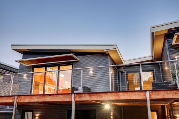 Exterior view of a contemporary multi-level residence with a flat, projecting roofline, dark horizontal cladding, warm timber soffits, extensive glazing, and a raised timber balcony with slim metal balustrading. Warm interior lighting contrasts with the cool dusk sky.