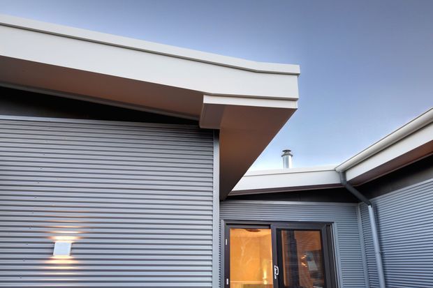 Exterior view of a contemporary low-profile building with silver-grey corrugated metal cladding, a large roller garage door, dark-framed glazed entry door, and projecting flat roof eaves surrounding a small courtyard-like junction.