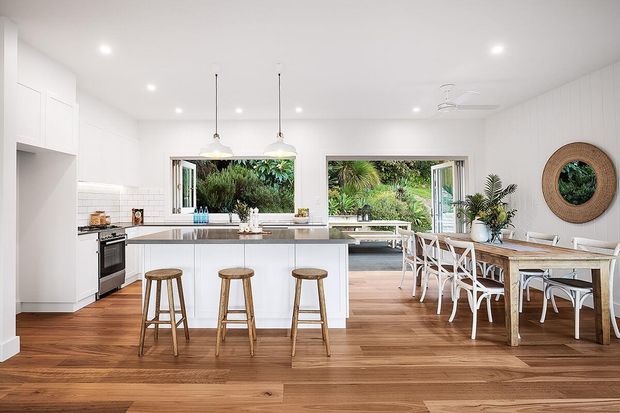 Bright open-plan kitchen and dining area with white cabinetry, a large island with three timber stools, pendant lighting, timber flooring, and a timber dining table with white chairs. Large openings connect the interior to lush greenery outside, while woven wall decor and indoor plants add a relaxed natural touch.