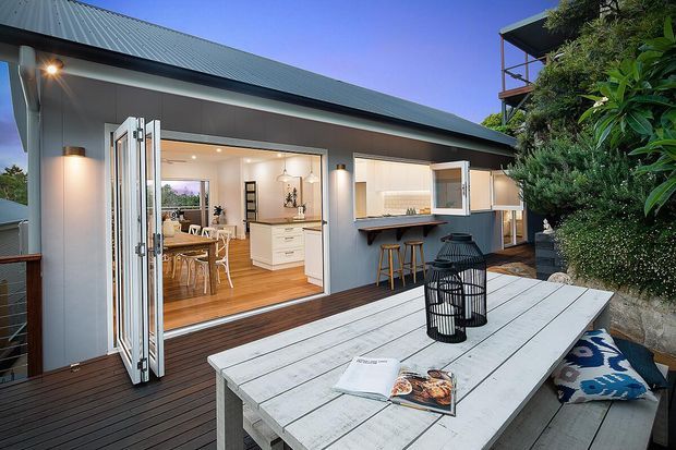 Exterior view of a contemporary home opening onto a timber deck and outdoor dining area. Wide bi-fold doors reveal a bright kitchen and dining space, while an adjacent outdoor servery with bar stools connects the interior to the deck. The building has a simple low-pitched metal roof, grey cladding, timber decking, and abundant surrounding greenery.