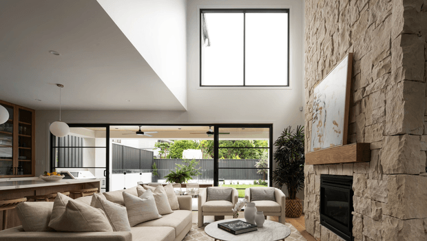 A bright, contemporary open-plan living room with a large cream sectional sofa, pale upholstered armchairs, a round light stone coffee table, and extensive glazing opening to a landscaped outdoor area. A substantial rough-stone fireplace wall with a timber mantel and inset fire forms the main feature, while a timber kitchen and built-in shelving are visible to the left. The room has a high ceiling with clerestory glazing, black-framed windows and doors, and a restrained neutral palette.