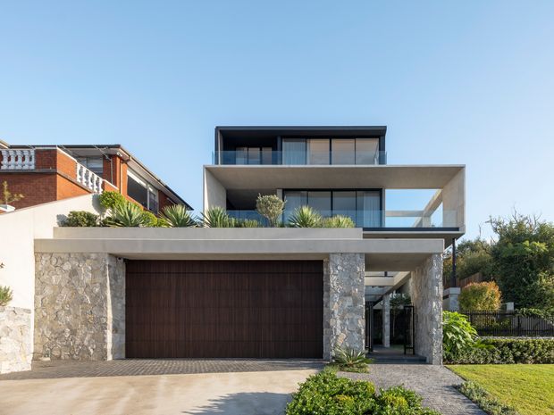 Front exterior view of a contemporary multi-level residence with strong horizontal concrete slab forms, expansive dark-framed glazing, a large timber-toned garage door, and rough natural-stone cladding at the lower level. Landscaped planting, a paved driveway, balcony areas, and a covered entry are visible. A neighbouring traditional brick home is partially visible to the left.