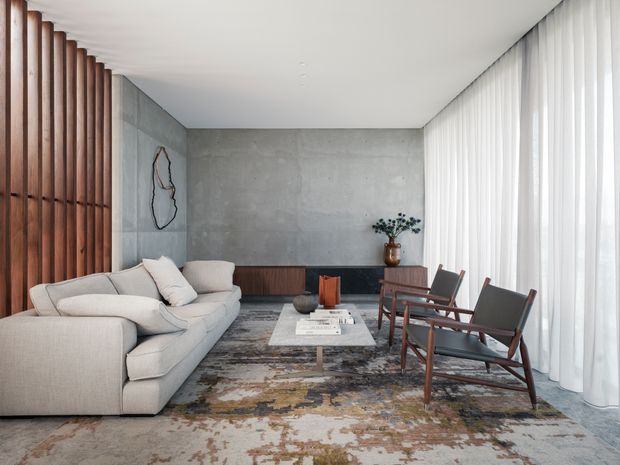 A refined contemporary living room with a large pale sectional sofa, two timber-framed lounge chairs, a low marble or stone coffee table, and a muted abstract rug. The room features a raw grey concrete feature wall, dark timber built-in cabinetry, a vertical timber slat screen, and full-height sheer curtains providing soft natural light. Minimal artwork and restrained decor create a calm, sophisticated atmosphere.