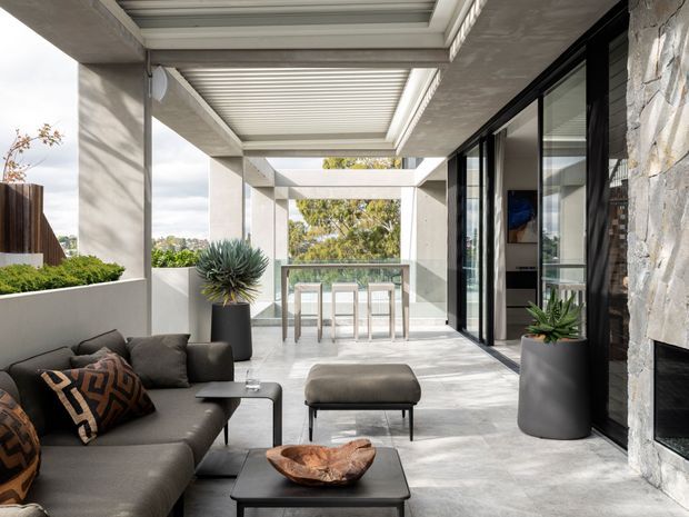 Contemporary covered outdoor living terrace with a grey sectional sofa, coffee table, ottoman, sculptural planter, and dining counter beyond. The space features large-format pale stone or concrete flooring, substantial rendered concrete columns and beams, a retractable louvred roof, frameless glass balustrade, black-framed sliding glazing, and a textured natural-stone fireplace wall. The terrace is bright, open, and connected to an interior room through the glazing.