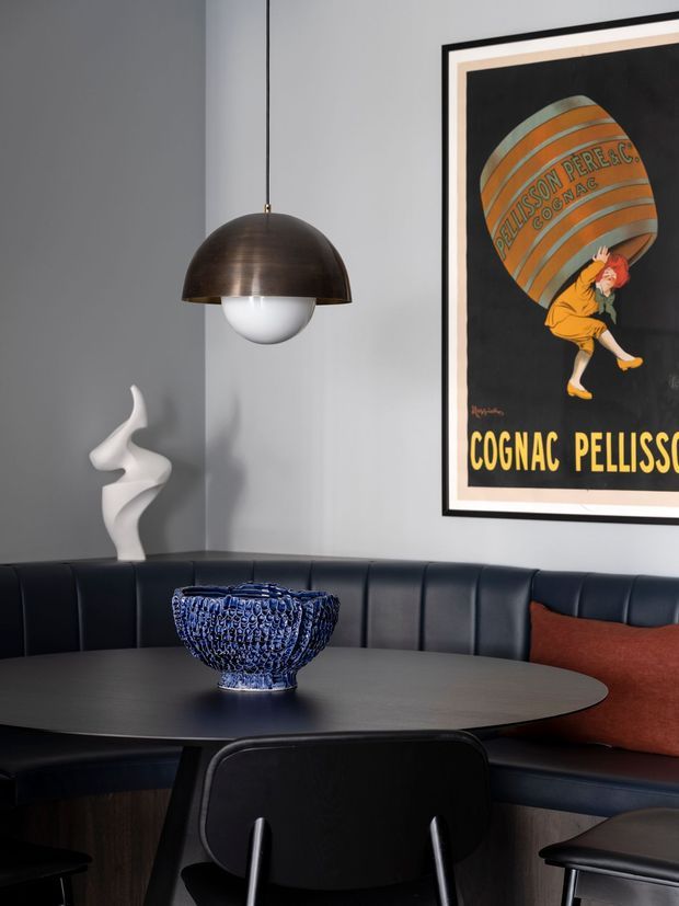 A moody, refined dining nook with a curved dark upholstered banquette wrapping around a round dark table. A black pendant with a bronze-toned dome and white globe hangs above the table, complemented by a blue glass bowl, white sculptural ornament, and a framed vintage cognac poster. The space uses dark navy-black seating, charcoal walls, and a rust-coloured cushion for contrast.