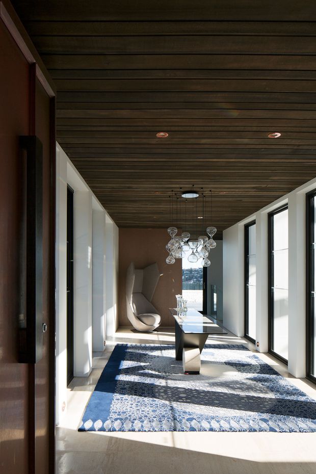 A long, narrow contemporary interior corridor or landing with full-height black-framed glazing along one side, white walls, a dark timber-lined ceiling with recessed downlights, and pale flooring. A sculptural black console table sits beneath a clustered glass pendant light, with a modern lounge chair at the far end. Strong sunlight creates bold shadows across a large blue-and-white patterned rug, giving the space a refined, gallery-like character.