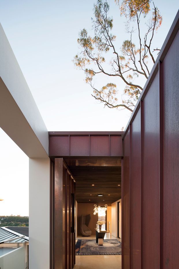 Architectural view toward a covered entrance or passage framed by a white projecting volume and dark reddish-brown timber or metal wall panels. The opening reveals a warmly lit interior hall with dark timber ceiling, patterned rug, sculptural seating, console furniture and a prominent decorative pendant light. A leafy tree and distant roofline are visible beyond, emphasizing the indoor-outdoor connection and courtyard-like approach.