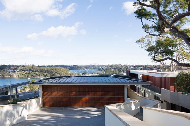 A modern residential exterior viewed from an elevated driveway or terrace, featuring a low-pitched dark metal roof, warm timber-clad garage door, white rendered walls, and adjoining contemporary forms. A broad harbour or waterfront landscape is visible beyond the property, with mature trees framing the scene.