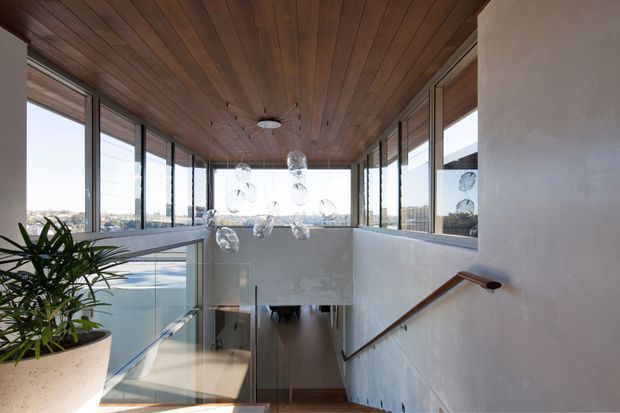 A bright contemporary stair landing with a descending timber stair and wooden handrail, enclosed by extensive floor-to-ceiling glazing. The space features a warm timber-lined ceiling, white rendered walls, a glass balustrade, and a cluster of clear pendant lights overlooking the lower level. A large potted plant sits at the landing edge, with an exterior view visible through the windows.