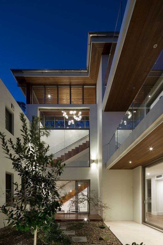 A dusk view into a narrow landscaped courtyard framed by a multi-level contemporary residence. White rendered walls, extensive glazing, glass balustrades, warm timber soffits and exposed interior staircases create a strong vertical architectural composition. Integrated lighting glows throughout the courtyard and interiors, with a small planted tree and gravel garden in the foreground.