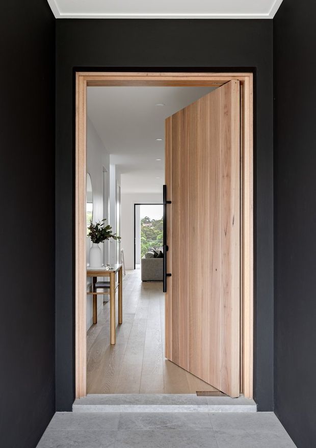 A dark, refined entryway viewed from outside a timber pivot front door. The doorway is framed in light timber against charcoal-black walls, with pale stone or concrete flooring at the threshold. Beyond the open door is a bright, minimalist hallway with light timber flooring, a narrow console table, vase, artwork, and views toward a living area and greenery outside.