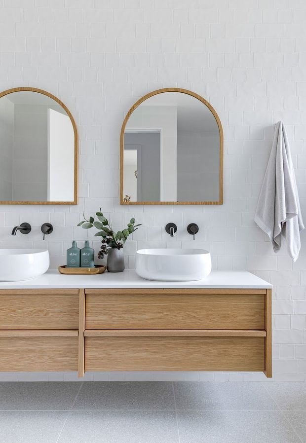 A bright, contemporary bathroom vanity with a double basin arrangement, wall-mounted light oak cabinetry, white vessel basins, matte black wall-mounted tapware, arched timber-framed mirrors, white textured wall tiles, and a light grey floor. A folded white towel hangs on the wall, while a small plant and toiletries sit on the vanity.