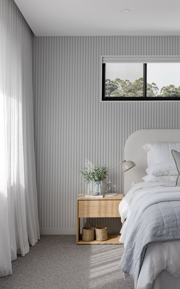 A bright, softly styled bedroom featuring a bed with a rounded upholstered headboard, layered white and pale grey bedding, a light timber bedside table, woven baskets, and a brass-toned reading lamp. The room has a full-height vertically fluted grey wall finish, sheer curtains, grey carpet, and a horizontal black-framed window overlooking trees.