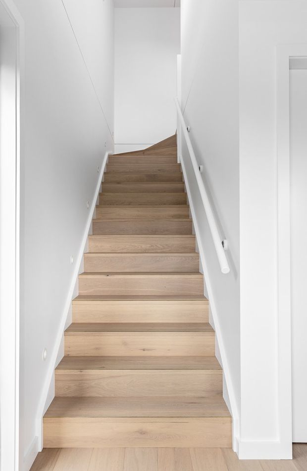 Minimal, bright interior photograph looking up a narrow straight staircase. The stair treads and risers are finished in light natural timber, set between smooth white walls with white skirting, trim, and a simple white handrail. A small landing and turn are visible at the top, with recessed wall lights and very limited decoration.