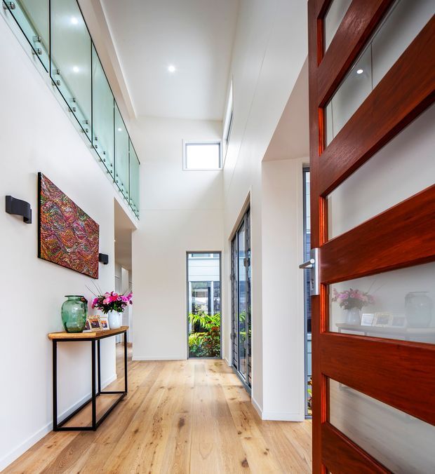 Bright contemporary entry hall with pale timber flooring, white walls, high ceilings, a glass balustrade overlooking an upper level, and extensive glazing at the far end. A large reddish-brown timber pivot door with inset glass panels dominates the foreground, while a slim console table and artwork decorate the hallway.