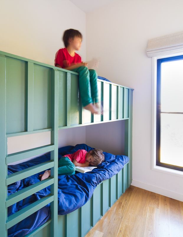 A bright children's bedroom featuring a custom-built green-painted bunk bed with vertical slatted railings and integrated ladder. Two children are using the bunk beds, with vivid blue bedding and a colorful palette. The room has white walls, a large black-framed window with a light roller blind, and warm timber flooring.