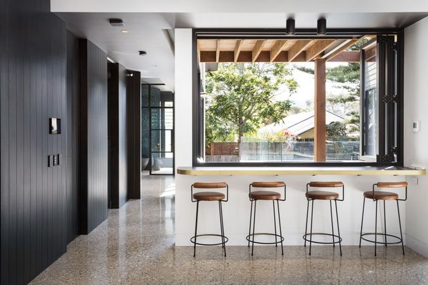 A bright contemporary kitchen and breakfast bar opening onto a covered outdoor area. The room features a long pale stone or engineered-stone counter with four slim bar stools, dark vertically grooved built-in cabinetry, polished terrazzo-style flooring, white walls and ceiling, and large black-framed opening windows. Timber roof beams and greenery are visible outside.