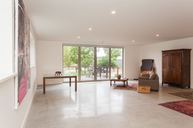 Bright, spacious minimalist living room with polished concrete flooring, white walls, recessed ceiling lights, and full-height sliding glass doors opening to an outdoor terrace. The room contains a simple timber console table, sculptural armchair, coffee table, vintage wooden armoire, woven basket, patterned rugs, and a large colourful wall artwork. Greenery and an outdoor seating area are visible beyond the glazing.
