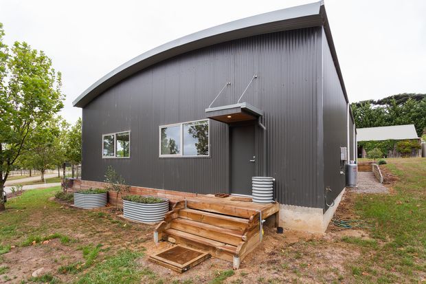 Exterior view of a compact dark-clad building with a distinctive curved gable roof, vertical ribbed metal wall cladding, simple rectangular windows, and a covered entry canopy reached by timber steps. The building sits in a landscaped suburban or rural setting with trees and lawn.