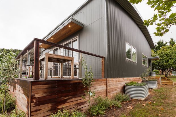 Exterior view of a contemporary single-storey building with a steep gable roof, dark vertical corrugated cladding, timber retaining walls, and a raised timber deck with wire balustrading. Large glazed doors open onto the deck, while landscaped planting and corrugated metal garden planters surround the structure.