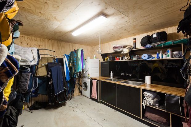 Compact utility and storage room lined with unfinished plywood, featuring extensive open shelving, black built-in cabinetry, a sink and benchtop, washing machine, drying racks, sports equipment, and wall-mounted storage. A bright linear fluorescent fixture illuminates the practical workshop-like space.
