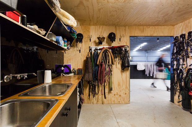 A compact utility and equipment room lined with plywood, featuring a stainless-steel double sink and timber benchtop, open shelving, and extensive wall-mounted storage for riding or outdoor gear. A doorway opens to a larger, brightly lit storage or work area beyond.