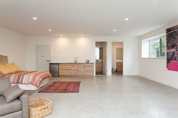 Bright, open-plan studio-style interior with a sleeping area, compact kitchenette, and adjoining bathroom visible through the doorway. The room has polished concrete flooring, white walls and ceiling, recessed lighting, light timber cabinetry, exposed white brick backsplash, and colourful textiles and artwork adding contrast.