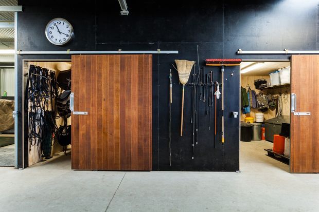 A utilitarian garage or workshop interior with a polished concrete floor and black wall panels. Two large timber sliding doors open onto organized storage closets containing outdoor and maintenance equipment, while tools and cleaning implements are mounted on the central wall. Industrial overhead lighting and exposed utility elements reinforce the functional workshop character.