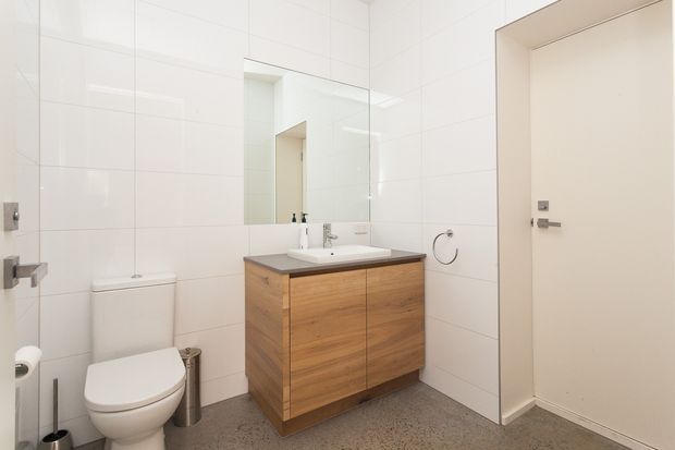 Bright contemporary bathroom with white tiled walls, a large wall mirror, a wall-mounted timber vanity with white basin, toilet, towel ring and stainless-steel fittings. The room has a polished grey concrete or terrazzo-style floor and a clean, functional layout.