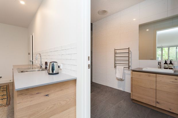 Bright contemporary interior showing a compact kitchenette on the left and a modern bathroom on the right. The kitchenette has a white benchtop, stainless-steel sink and tapware, white subway-tile splashback, and light timber cabinetry. The bathroom features large light wall tiles, dark grey floor tiles, a timber vanity with a white basin, illuminated mirror, and heated towel rail.