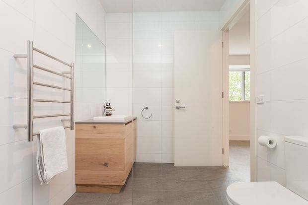 Bright, contemporary bathroom with large white wall tiles, grey floor tiles, a timber vanity with white basin, wall-mounted towel warmer, toilet and a frameless shower screen. An open doorway leads directly to a bedroom or adjacent sleeping area visible beyond.