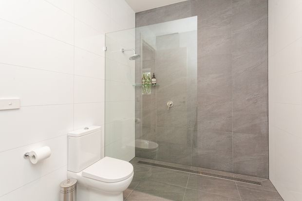 A clean, contemporary bathroom featuring a wall-mounted toilet, frameless glass walk-in shower, large-format light grey wall and floor tiles, a recessed shower shelf, and linear floor drainage. White tiled walls and streamlined fixtures create a bright, minimalist feel.