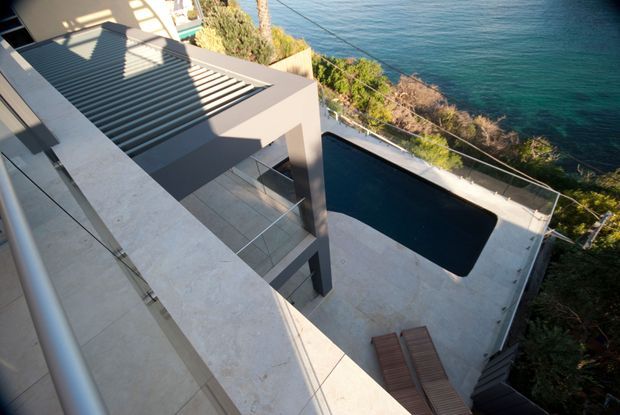 Elevated view of a modern multi-level coastal residence with a flat roof and shaded pergola, broad pale stone or concrete terraces, and a dark rectangular swimming pool overlooking the ocean. The image emphasizes the building’s geometric forms, outdoor entertaining levels, and dramatic waterfront setting.
