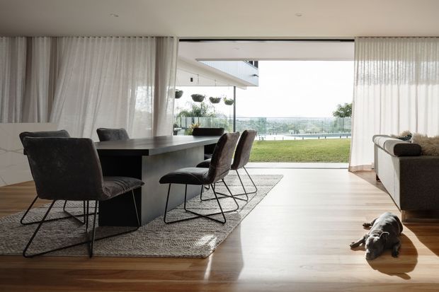 A refined open-plan dining and living area with a dark rectangular dining table, upholstered dining chairs, a textured rug, and a neutral sofa. Full-height sheer curtains frame wide glazing and an open connection to a lawn and distant landscape. Light timber flooring, a restrained neutral palette, and abundant natural light create a calm contemporary setting.