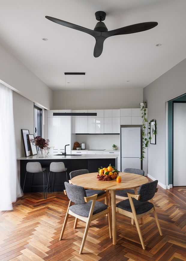 Bright open-plan kitchen and dining area with a round timber dining table and four upholstered timber-framed chairs in the foreground. The kitchen has white flat-panel cabinetry, a white island with black base cabinetry, integrated appliances, black pendant lighting, and a dark sculptural ceiling fan. Herringbone timber flooring, white walls, large windows with sheer curtains, indoor plants, and understated modern furnishings create a clean, warm contemporary interior.