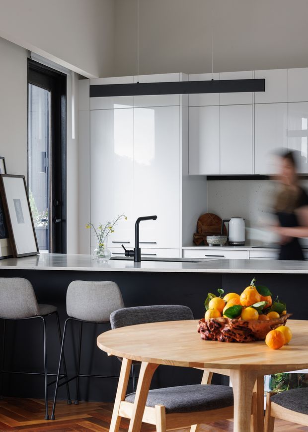 A bright contemporary kitchen and dining area with glossy white full-height cabinetry, a long black linear pendant light, and a dark kitchen island topped with a pale stone benchtop. A matte black tap and sink sit at the island, with upholstered bar stools facing the kitchen. In the foreground, a round light-wood dining table and chairs occupy the timber-floored space. A tall black-framed glazed door or window brings in natural light, while a fruit bowl and simple vase provide restrained styling.