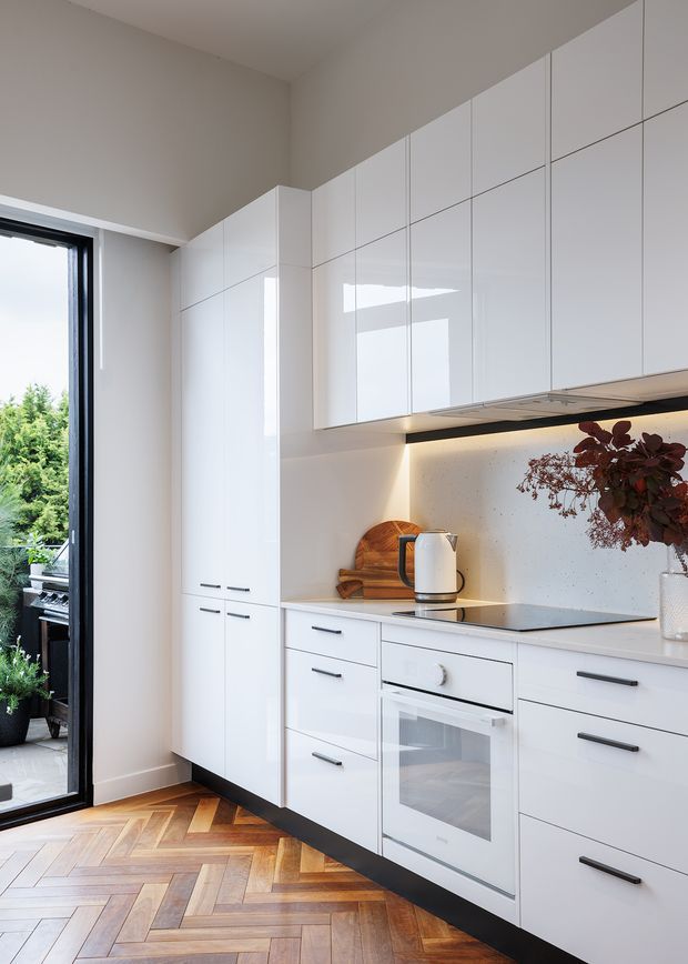 A compact, contemporary white kitchen with full-height glossy white cabinetry, integrated appliances, a pale speckled backsplash, and under-cabinet lighting. The kitchen has a warm herringbone timber floor, dark linear hardware and plinth, and a black-framed glazed door opening toward greenery. A timber cutting board, kettle, and dried foliage provide restrained decorative accents.