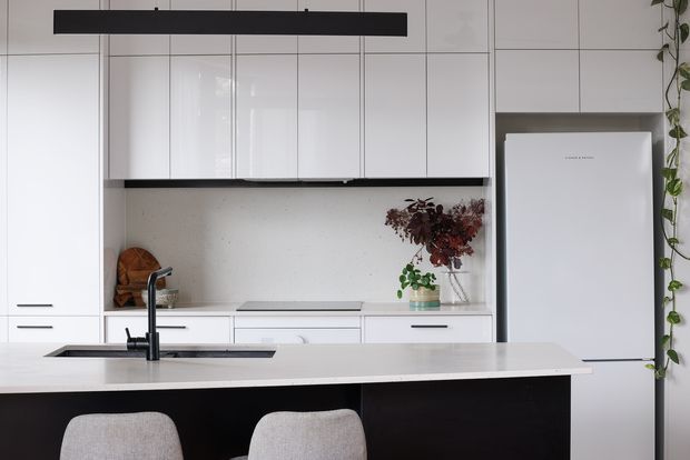 A bright, contemporary kitchen viewed straight on, with a large pale stone island and integrated sink, black island cabinetry, glossy white full-height cabinetry, a white refrigerator, and a light speckled backsplash. A slim black linear pendant provides contrast, while restrained greenery and dried foliage add a natural accent.