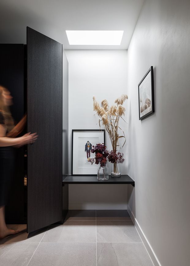 A narrow, contemporary entry hall with pale grey tiled flooring, smooth light walls, and a dark timber pivot door opening to the left. A slim black console shelf displays framed artwork and dried floral arrangements, while a rectangular skylight brightens the corridor. A person is partially visible behind the door.
