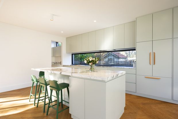 Bright contemporary kitchen with a large central island, integrated pale cabinetry, white stone benchtops, and a long horizontal window. Green bar stools add contrast, while the timber floor brings warmth to the clean, light-filled space.