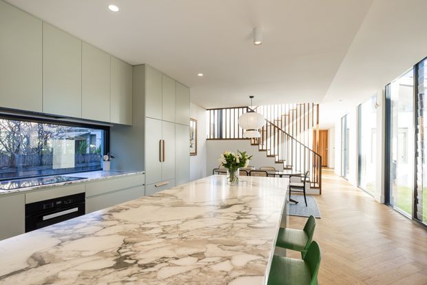 Bright contemporary open-plan kitchen and dining area with a large veined stone island, pale green built-in cabinetry, integrated appliances, green bar stools, and a timber herringbone floor. A sculptural pendant hangs above the dining table, while a timber stair with vertical slatted balustrade and extensive glazing extends along the right side of the space.
