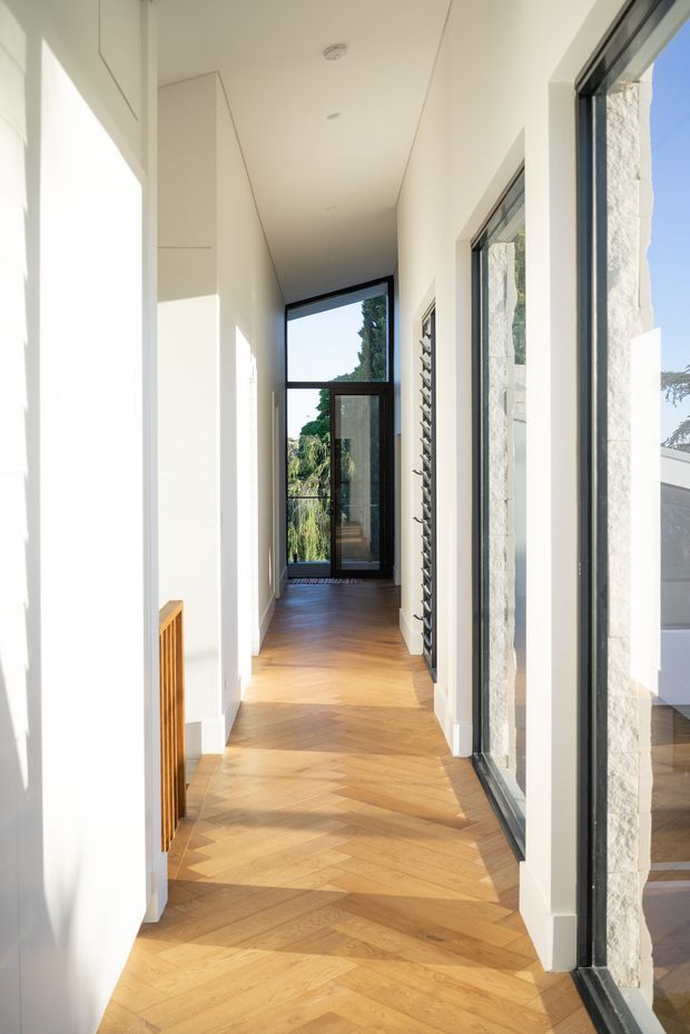 A bright, narrow residential hallway with white walls and concealed storage, extensive black-framed glazing, and a warm timber herringbone floor. Natural light floods the corridor, with views into greenery and a textured pale stone wall visible through the windows. A small timber wall feature or radiator cover appears along the left side.