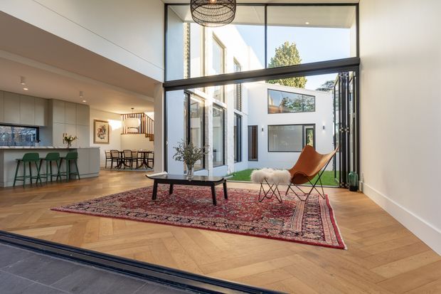 A bright, double-height contemporary living area opening onto an internal courtyard through expansive black-framed glazing and folding doors. The room features warm herringbone timber flooring, a large patterned red rug, a low coffee table, an orange lounge chair, and a small wire-frame side table. A kitchen with a pale island and green bar stools, along with a dining area, is visible to the left. A sculptural pendant hangs above the space, and the courtyard-facing architecture includes white walls, tall windows, and a small patch of lawn.