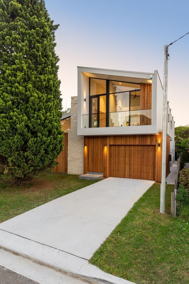 Street-facing view of a contemporary multi-storey residence at dusk, with a white rendered rectangular upper volume, extensive black-framed glazing, a glass balcony balustrade, warm timber vertical screening and a timber garage door. A pale stone-clad wall, concrete driveway, lawn, mature evergreen tree and utility pole frame the facade.