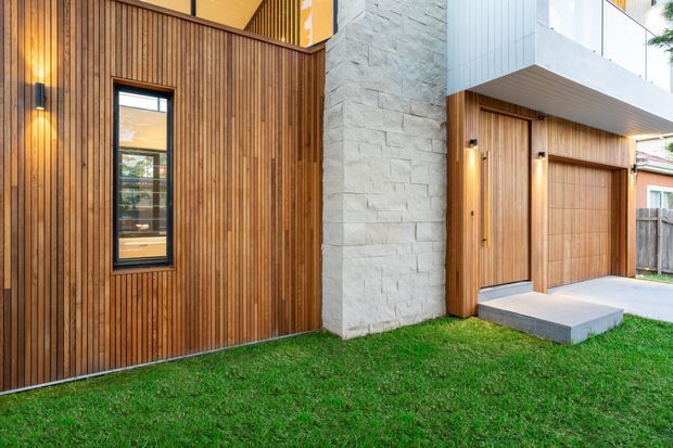 A contemporary residential facade viewed from the front, featuring vertical timber cladding, a tall light-grey textured masonry feature wall, dark-framed glazing, and an integrated timber garage door beneath a projecting upper-level volume. A simple concrete entry step and lawn complete the scene.