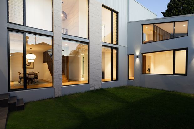 A modern multi-level residential building arranged around a small grass courtyard. The facade combines pale rendered and textured masonry walls with large black-framed windows and glazed openings, revealing a warm, softly lit dining area, staircase, and interior spaces. The composition highlights the courtyard-facing architecture at dusk, with clean geometric forms and strong indoor-outdoor visual connections.