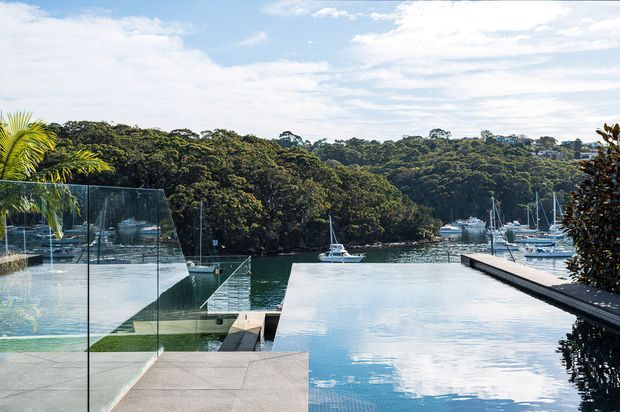 A contemporary waterfront pool terrace overlooking a sheltered marina and densely wooded shoreline. The image features a long reflective swimming pool, glass balustrades, pale stone or concrete paving, dark built-in pool edging, tropical planting, and moored boats in the background under a bright partly cloudy sky.