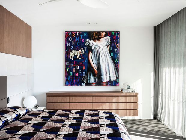 Bright contemporary bedroom with a low-profile walnut-toned dresser, white walls, floor-to-ceiling sheer curtains, and a bold colorful artwork above the dresser. The foreground features a patterned quilted bedspread in navy, black, white, and blush tones. Built-in wall cabinetry and a minimalist ceiling contribute to the clean, modern fit-out.