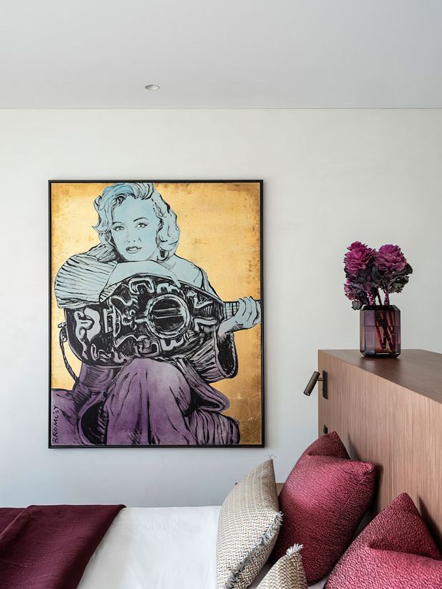 A close-up view of a contemporary bedroom with a white wall, recessed ceiling light, large framed pop-art artwork above the bed, and a timber-toned built-in bedside cabinet. The bed is dressed in white linen with burgundy, mauve, and textured neutral cushions, while a dark glass vase with deep purple flowers sits on the cabinet.